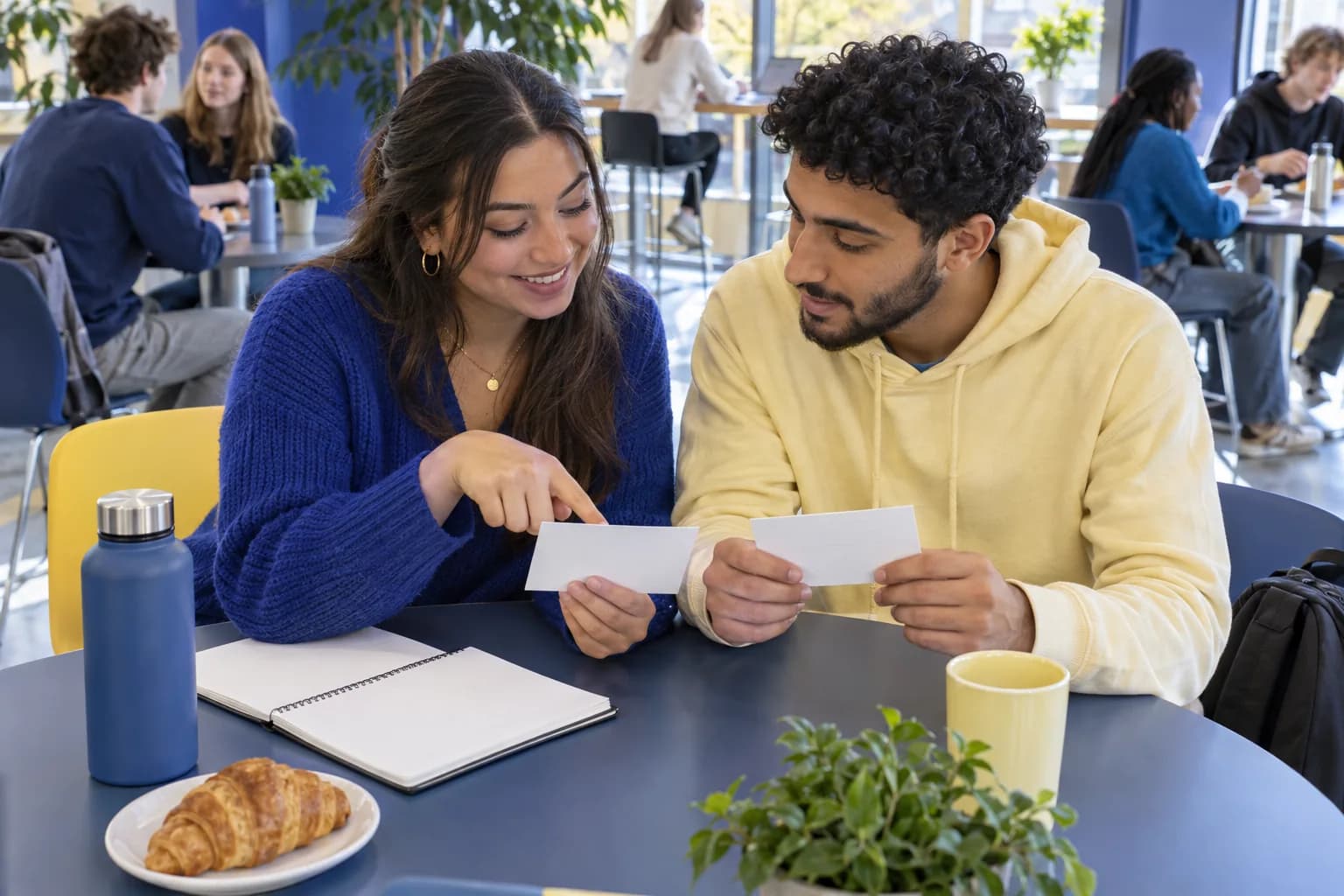 Two students discussing blank practice cards at a university cafeteria table